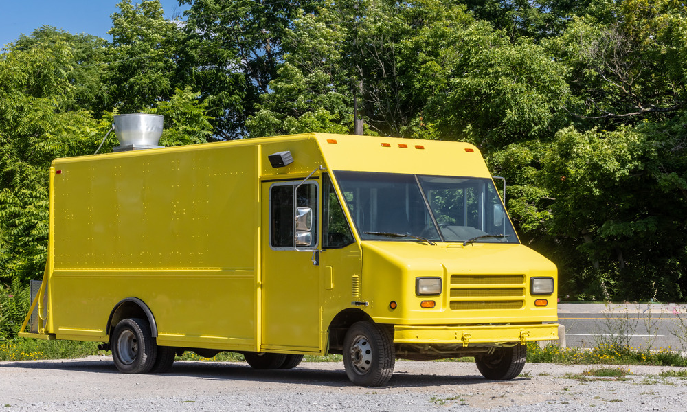 An empty yellow stepvan is parked outside in front of a beautiful forest and an asphalt road on a sunny day.