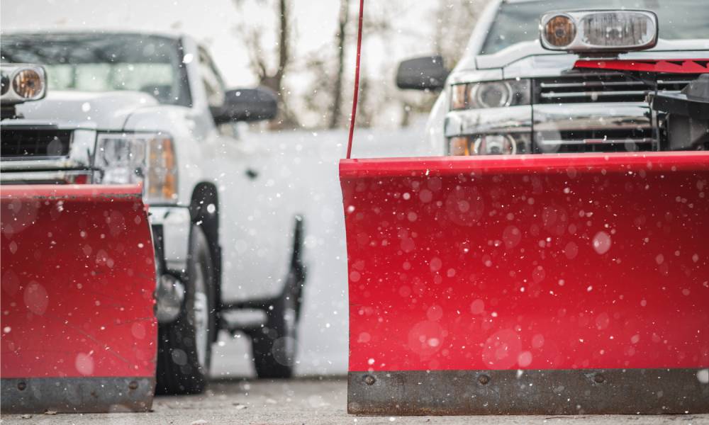 Two trucks equipped with snow plows that have red blades are parked outside on a snowy day, and nobody is nearby.